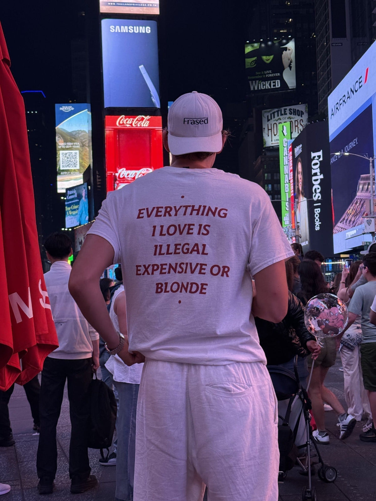 Person wearing a white FRASED Everything I Love Is Illegal Expensive or Blonde tee in Times Square at night