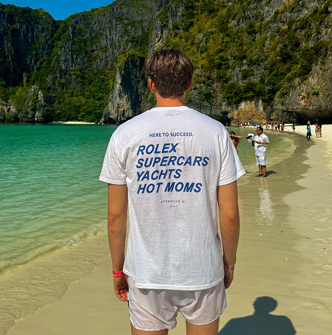 Person wearing a white FRASED Here to Succeed T-shirt with blue Rolex Supercars Yachts Hot Moms print on a tropical beach