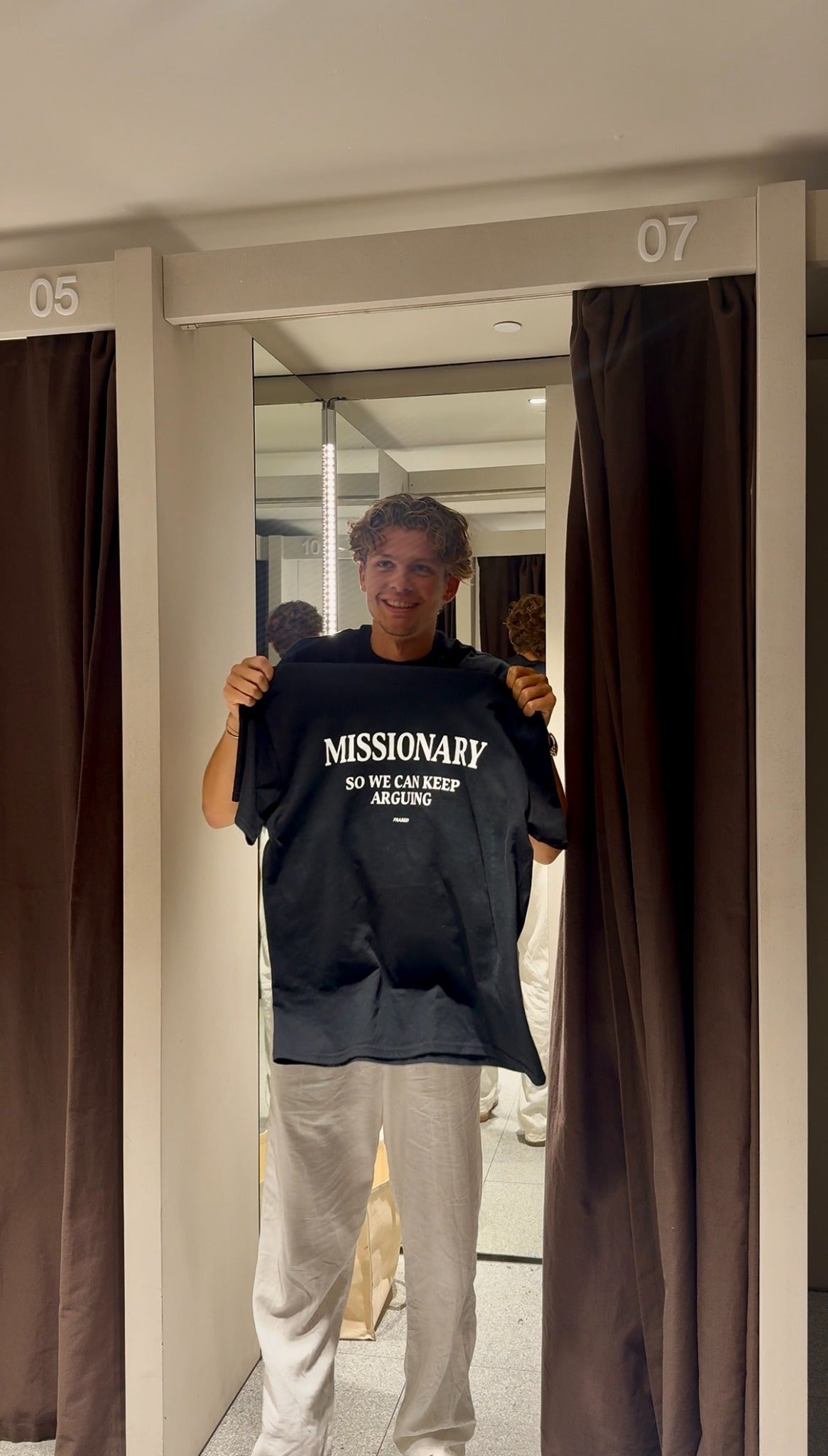 Person holding a black FRASED Missionary So We Can Keep Arguing tee in a fitting room