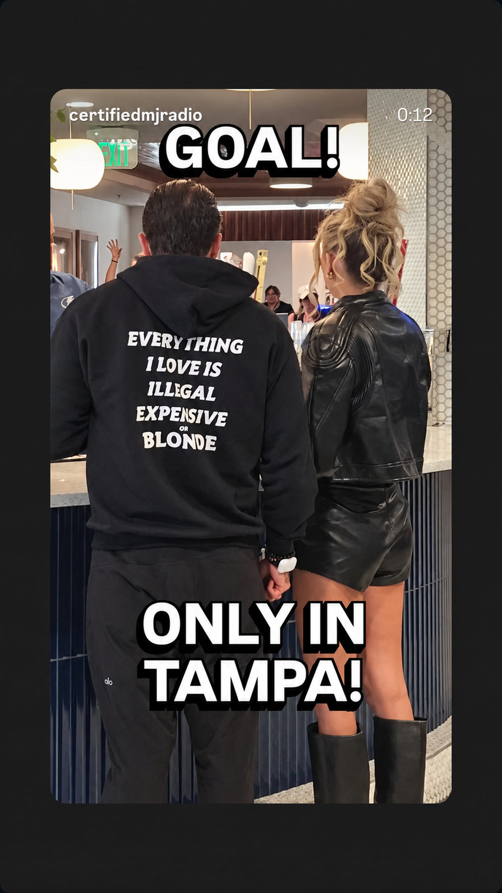 Man in a black “EVERYTHING I LOVE IS ILLEGAL, EXPENSIVE OR BLONDE” hoodie standing beside a woman.