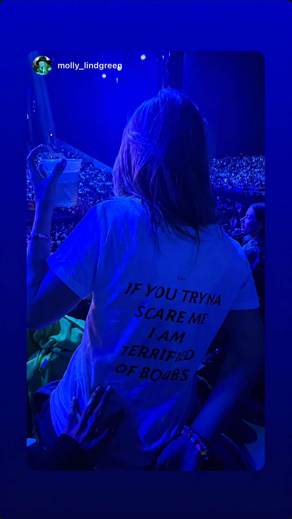Kvinde i hvid IF YOU TRYNA SCARE ME, I AM TERRIFIED OF BOOBS T-shirt blandt et koncertpublikum i blåt lys.