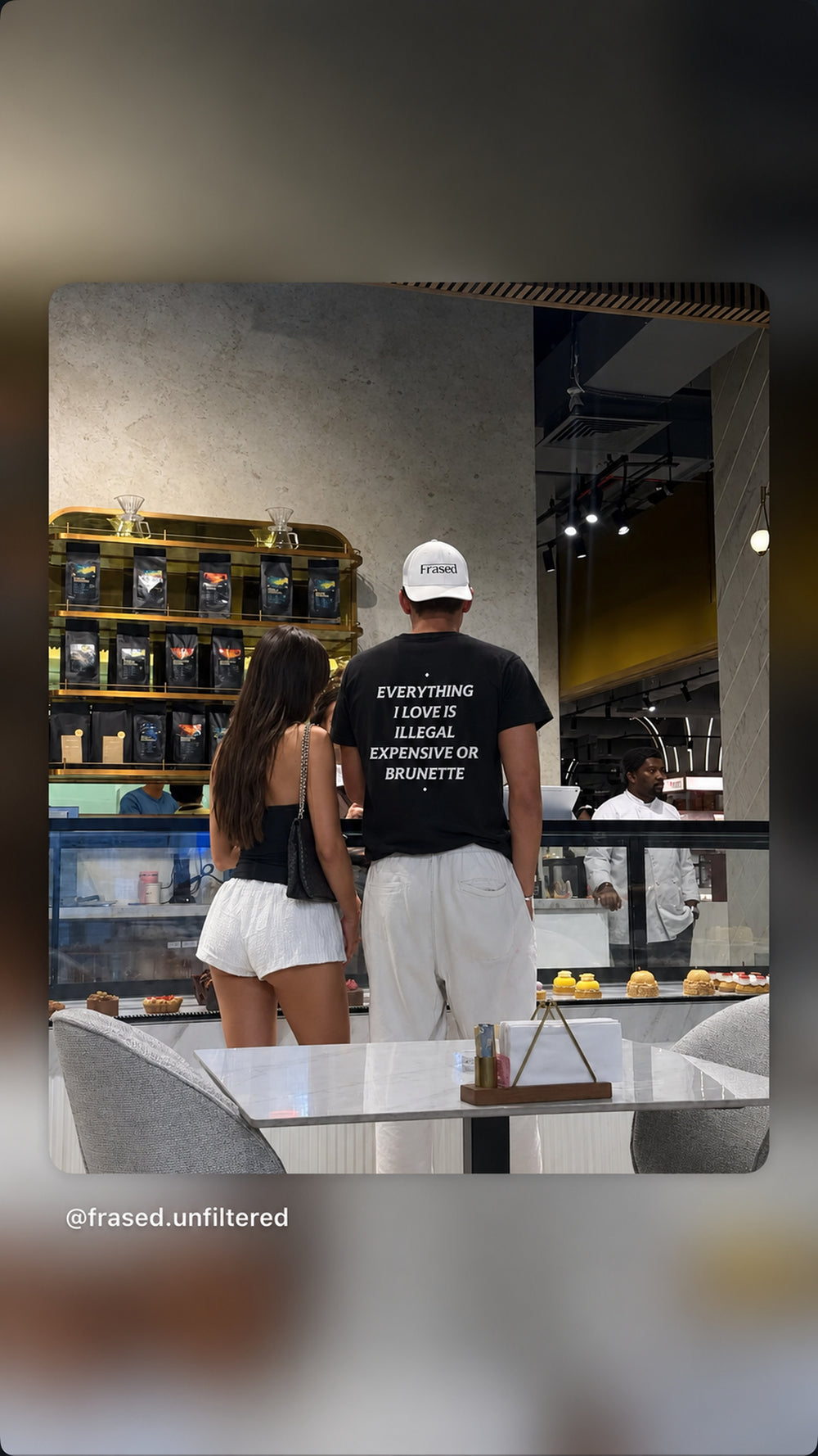 Mand i sort EVERYTHING I LOVE IS ILLEGAL, EXPENSIVE OR BRUNETTE T-shirt står med en kvinde ved en café.
