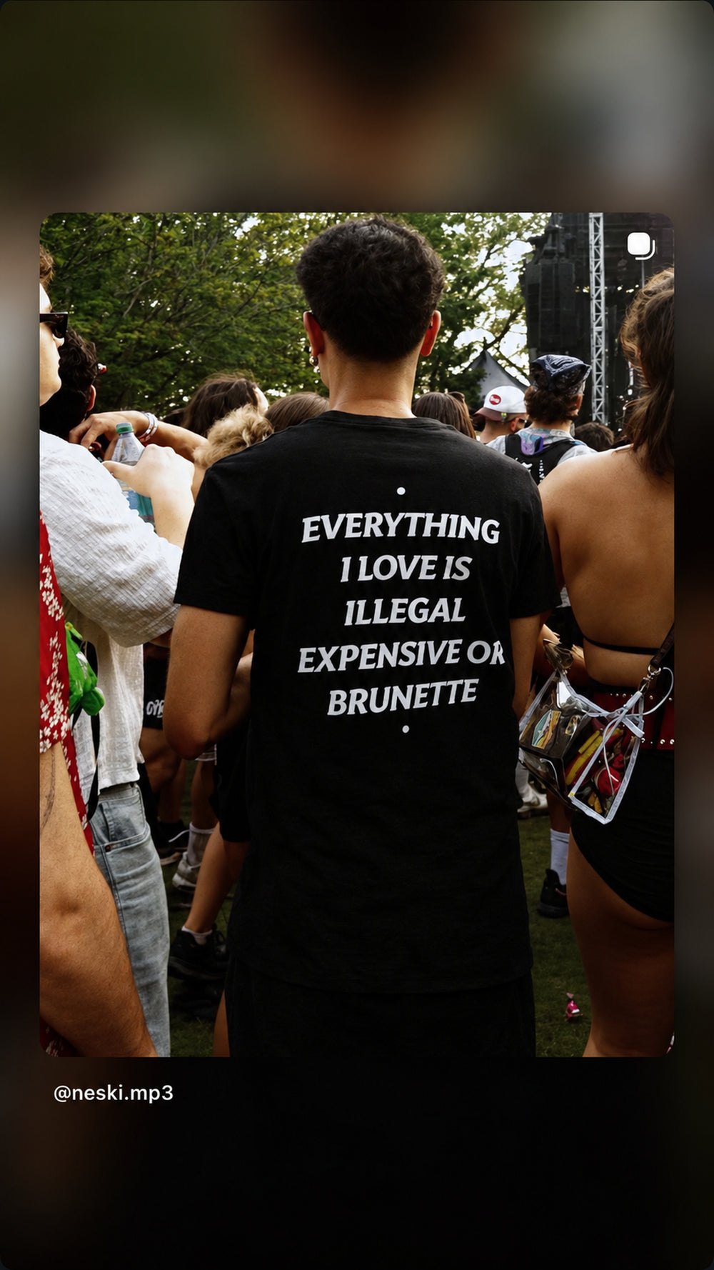 Person i sort EVERYTHING I LOVE IS ILLEGAL, EXPENSIVE OR BRUNETTE T-shirt blandt koncertgæster udendørs.