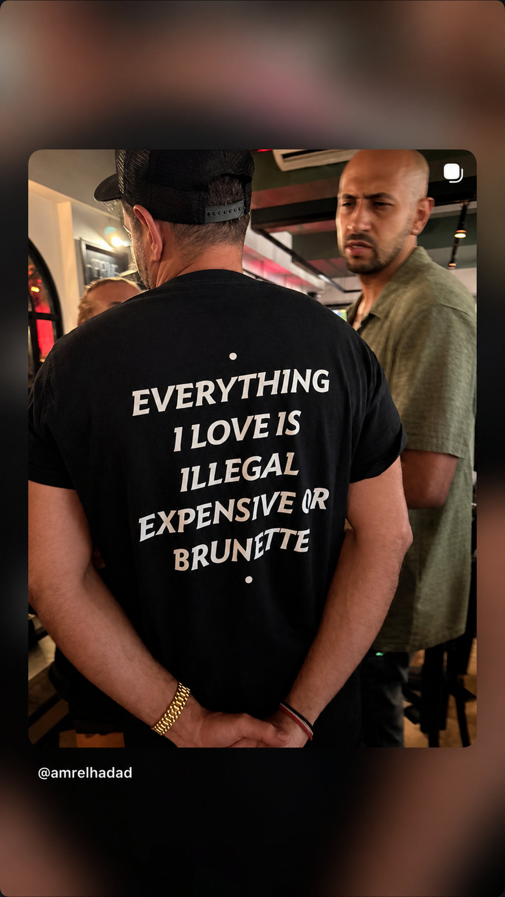 Mand i sort EVERYTHING I LOVE IS ILLEGAL, EXPENSIVE OR BRUNETTE T-shirt i en bar.