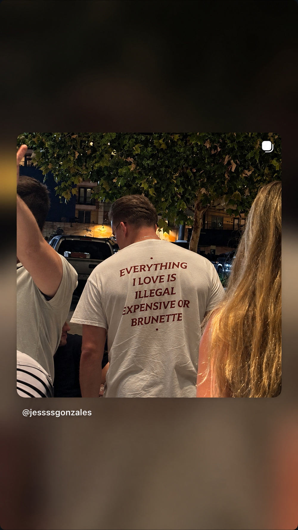 Person i hvid EVERYTHING I LOVE IS ILLEGAL, EXPENSIVE OR BRUNETTE T-shirt i en menneskemængde om aftenen.