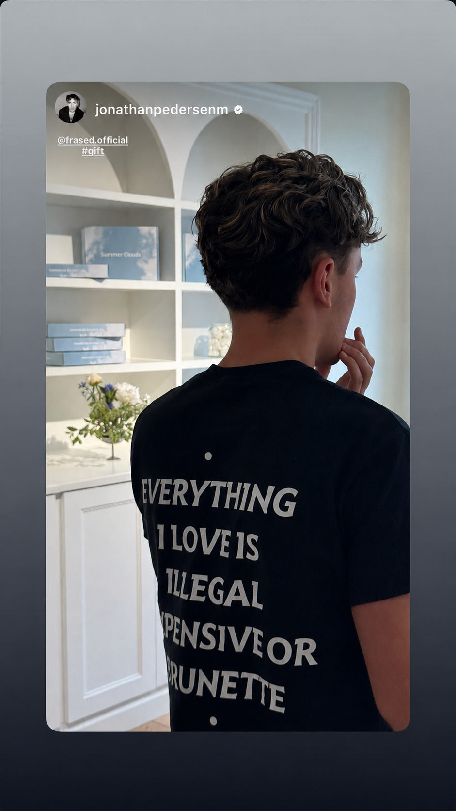 Mand i sort EVERYTHING I LOVE IS ILLEGAL, EXPENSIVE OR BRUNETTE T-shirt indendørs foran hvide hylder.