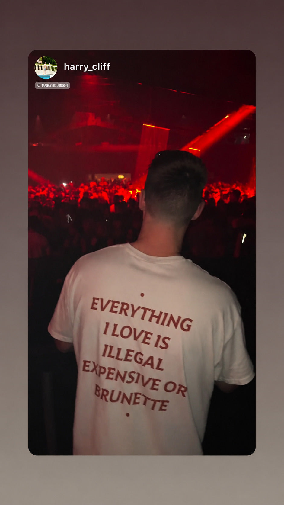 Mand i hvid EVERYTHING I LOVE IS ILLEGAL, EXPENSIVE OR BRUNETTE T-shirt foran et rødoplyst koncertpublikum.