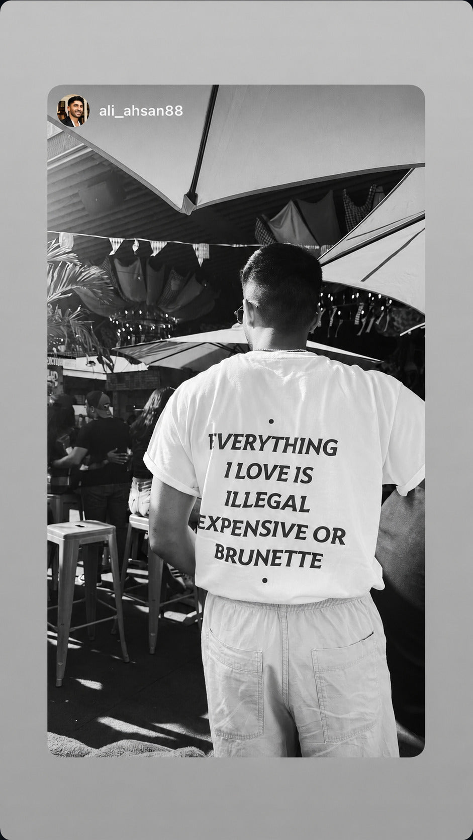 Sort-hvidt foto af mand i hvid EVERYTHING I LOVE IS ILLEGAL, EXPENSIVE OR BRUNETTE T-shirt på en terrasse.