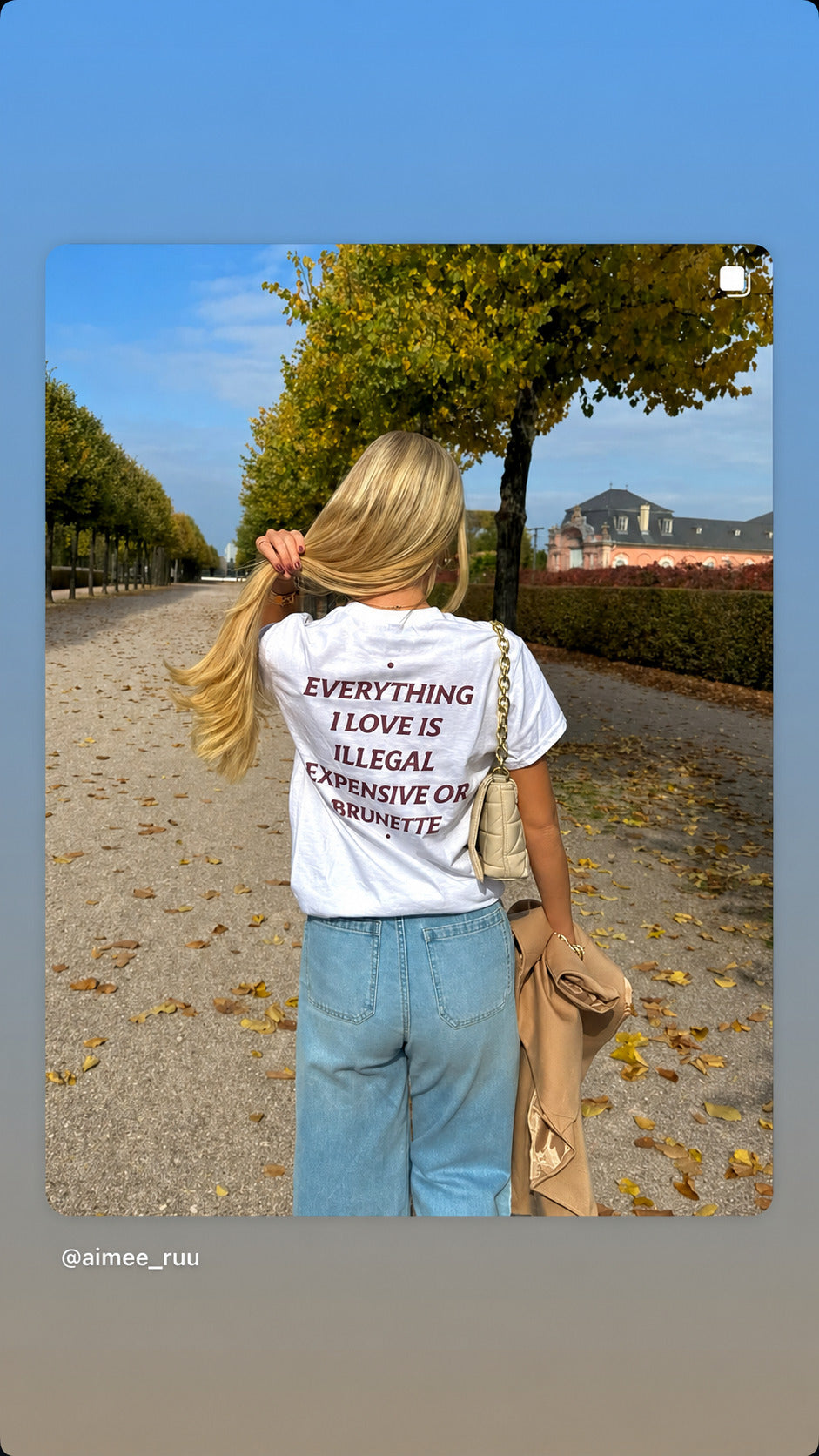 Kvinde i hvid EVERYTHING I LOVE IS ILLEGAL, EXPENSIVE OR BRUNETTE T-shirt og jeans på en træomkranset sti.