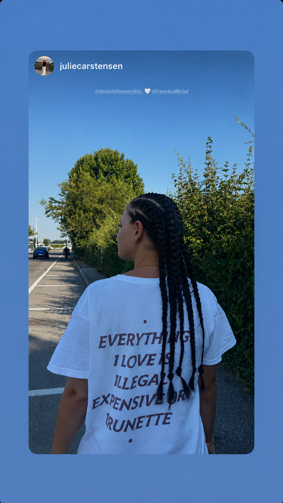 Kvinde med fletninger i hvid EVERYTHING I LOVE IS ILLEGAL, EXPENSIVE OR BRUNETTE T-shirt på en vej.