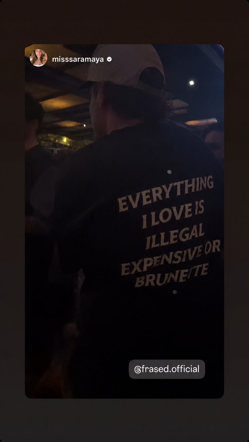 Person i sort EVERYTHING I LOVE IS ILLEGAL, EXPENSIVE OR BRUNETTE T-shirt i en mørk bar.