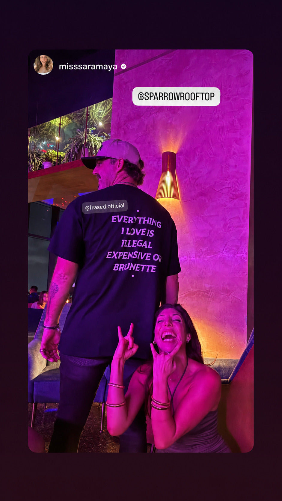 Mand i sort EVERYTHING I LOVE IS ILLEGAL, EXPENSIVE OR BRUNETTE T-shirt ved siden af en kvinde i lilla klublys.