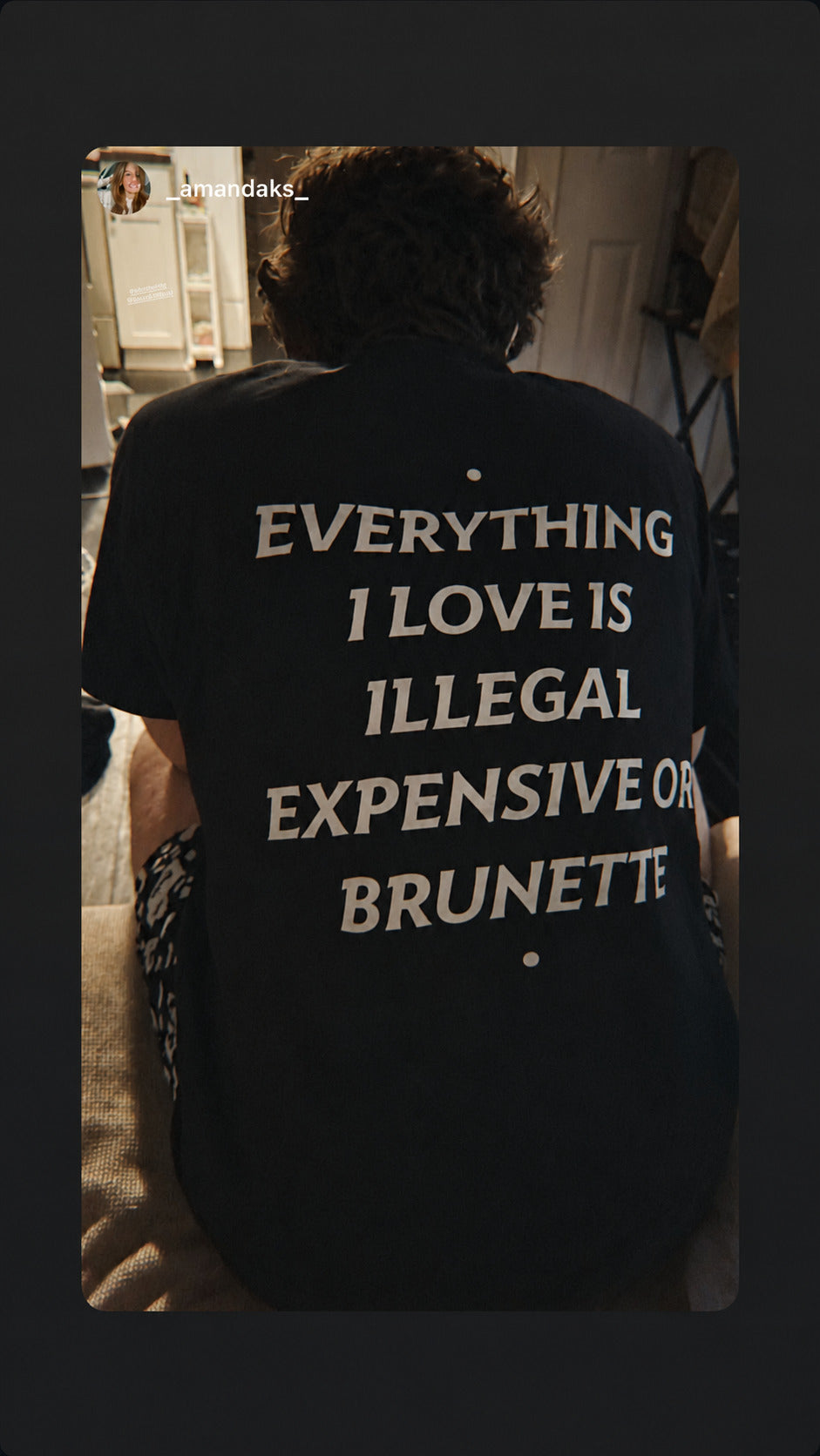 Nærbillede af sort EVERYTHING I LOVE IS ILLEGAL, EXPENSIVE OR BRUNETTE T-shirt på en person indendørs.