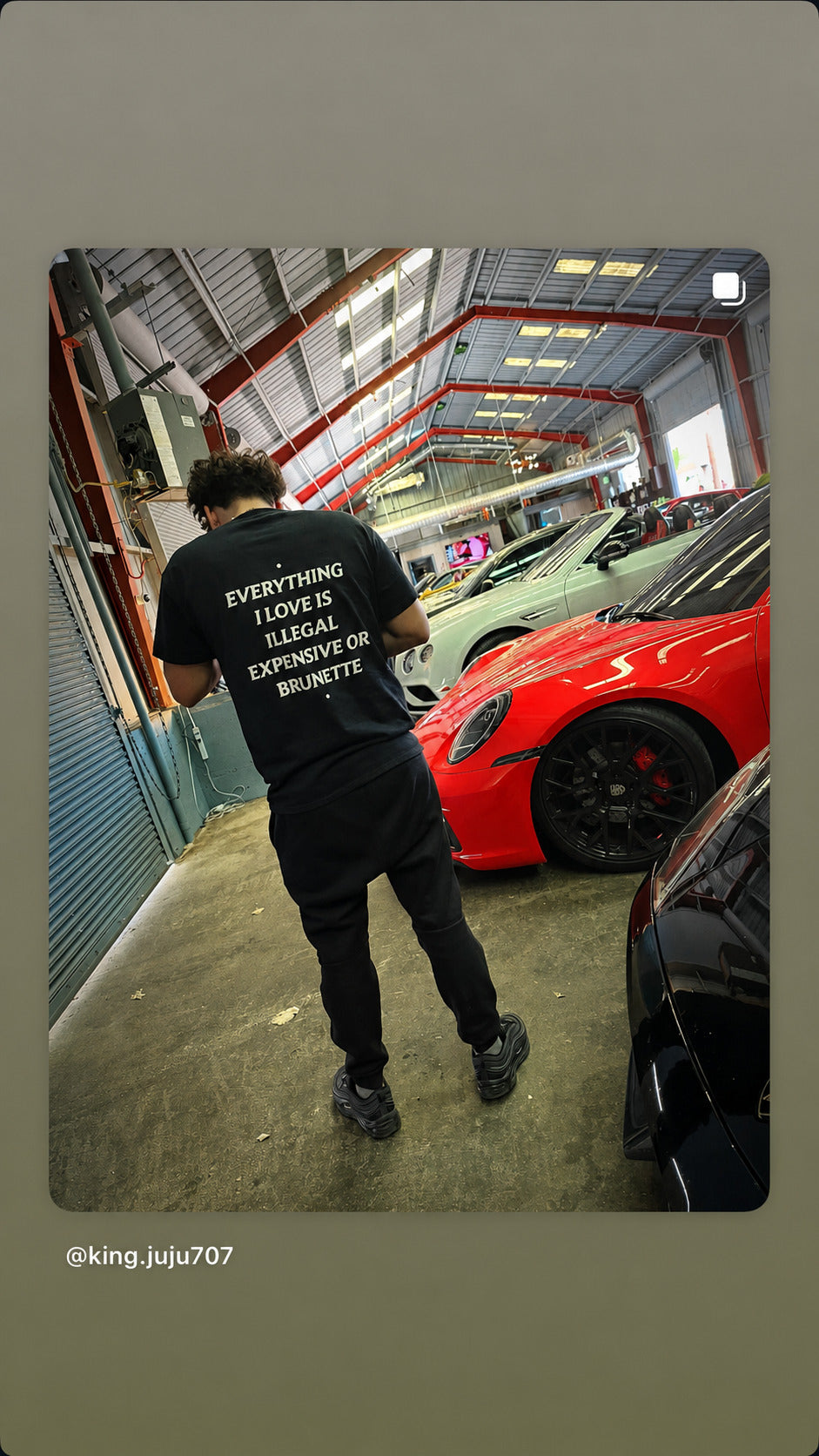 Mand i sort EVERYTHING I LOVE IS ILLEGAL, EXPENSIVE OR BRUNETTE T-shirt går mellem sportsvogne i en garage.