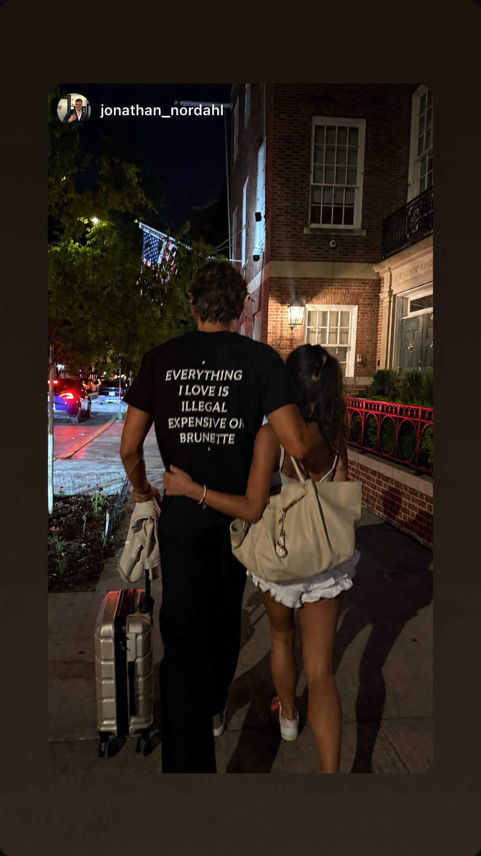 Mand i sort EVERYTHING I LOVE IS ILLEGAL, EXPENSIVE OR BRUNETTE T-shirt går arm i arm med en kvinde om aftenen.