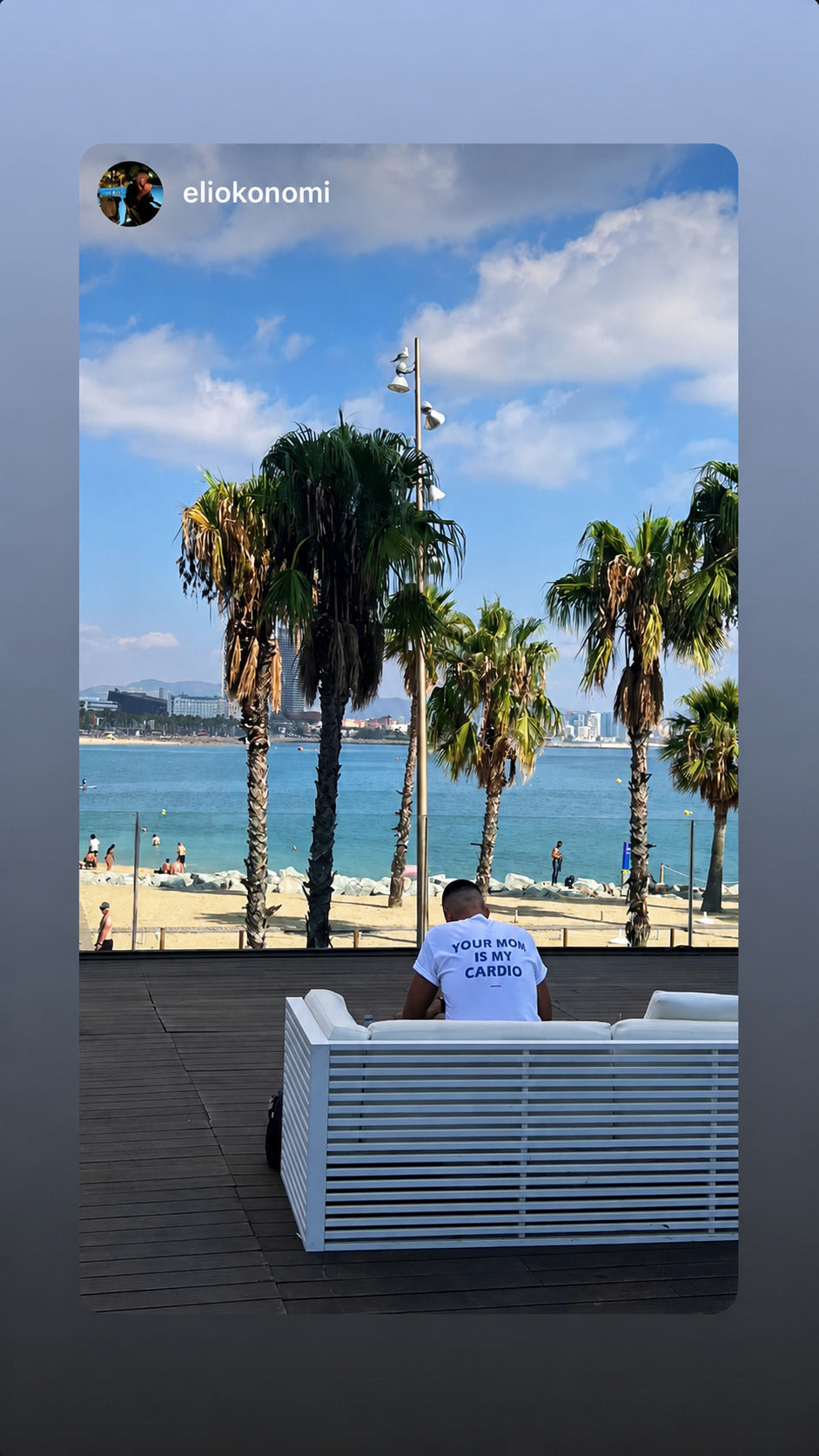 Person sidder ved stranden under palmer i hvid FRASED T-shirt med 'Your Mom Is My Cardio' på ryggen.