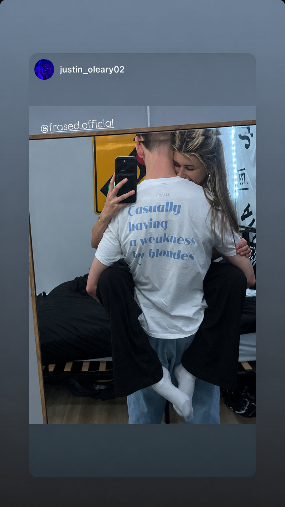 Par tager spejlbillede; mandens hvide FRASED T-shirt viser rygtrykket 'Casually having a weakness for blondes' i lyseblåt.