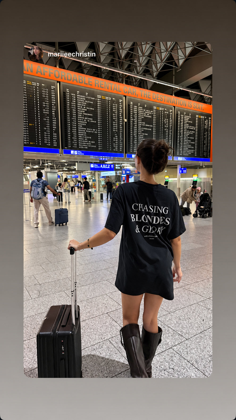 Traveler with a suitcase at an airport in a black FRASED T-shirt with “Chasing Blondes & Glory” on the back.