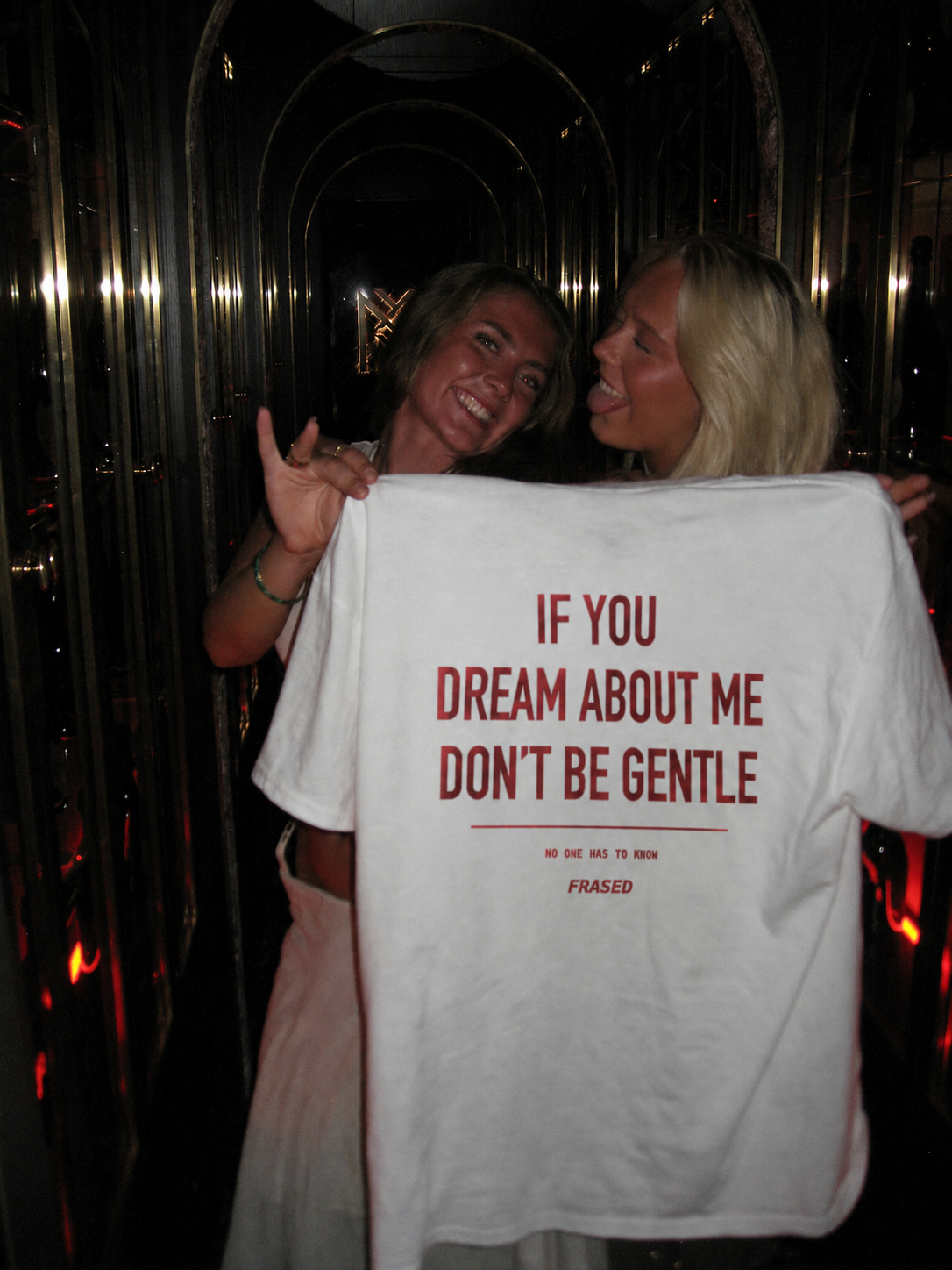 Two women posing in a dimly lit venue, holding up a white graphic t-shirt with red text that reads 'IF YOU DREAM ABOUT ME DON'T BE GENTLE' and 'FRANCED' underneath.