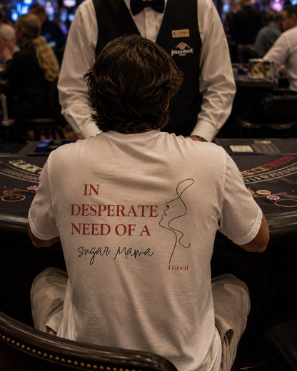 A person sitting at a blackjack table in a casino, viewed from behind, wearing a white t-shirt with red and black text that reads 'IN DESPERATE NEED OF A sugar Mama' and a small 'FRASED' logo, with a casino attendant standing in front.