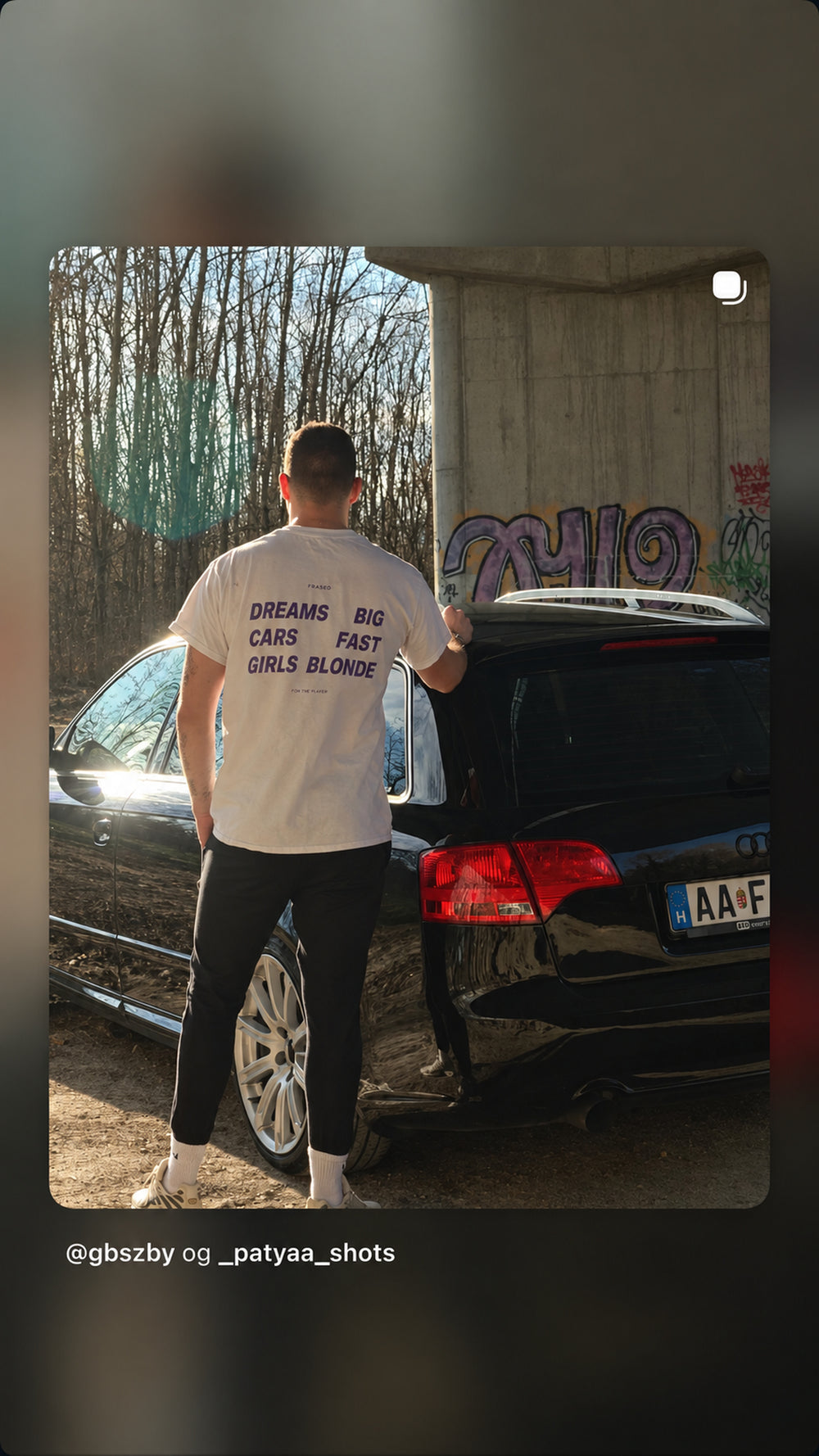 Mand ved sort bil iført hvid FRASED T-shirt med lilla 'Dreams Big. Cars Fast. Girls Blonde'-rygtryk.