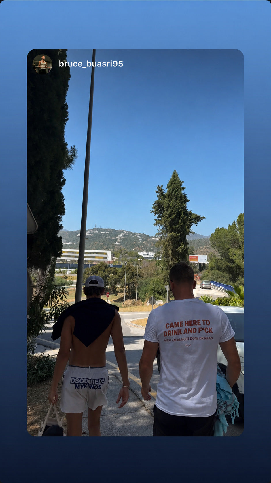 Two people walking on a sunny road; one wears a white “CAME HERE TO DRINK AND F*CK” T-shirt.