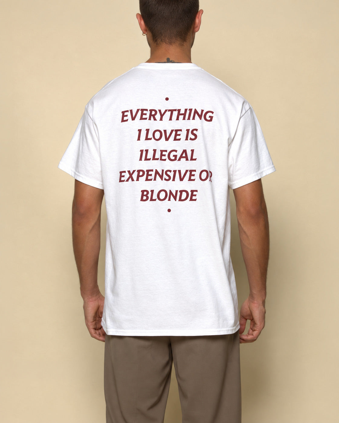 A man viewed from behind wearing a white t-shirt with a bold red text graphic on the back that reads 'EVERYTHING I LOVE IS ILLEGAL EXPENSIVE OR BLONDE'.
