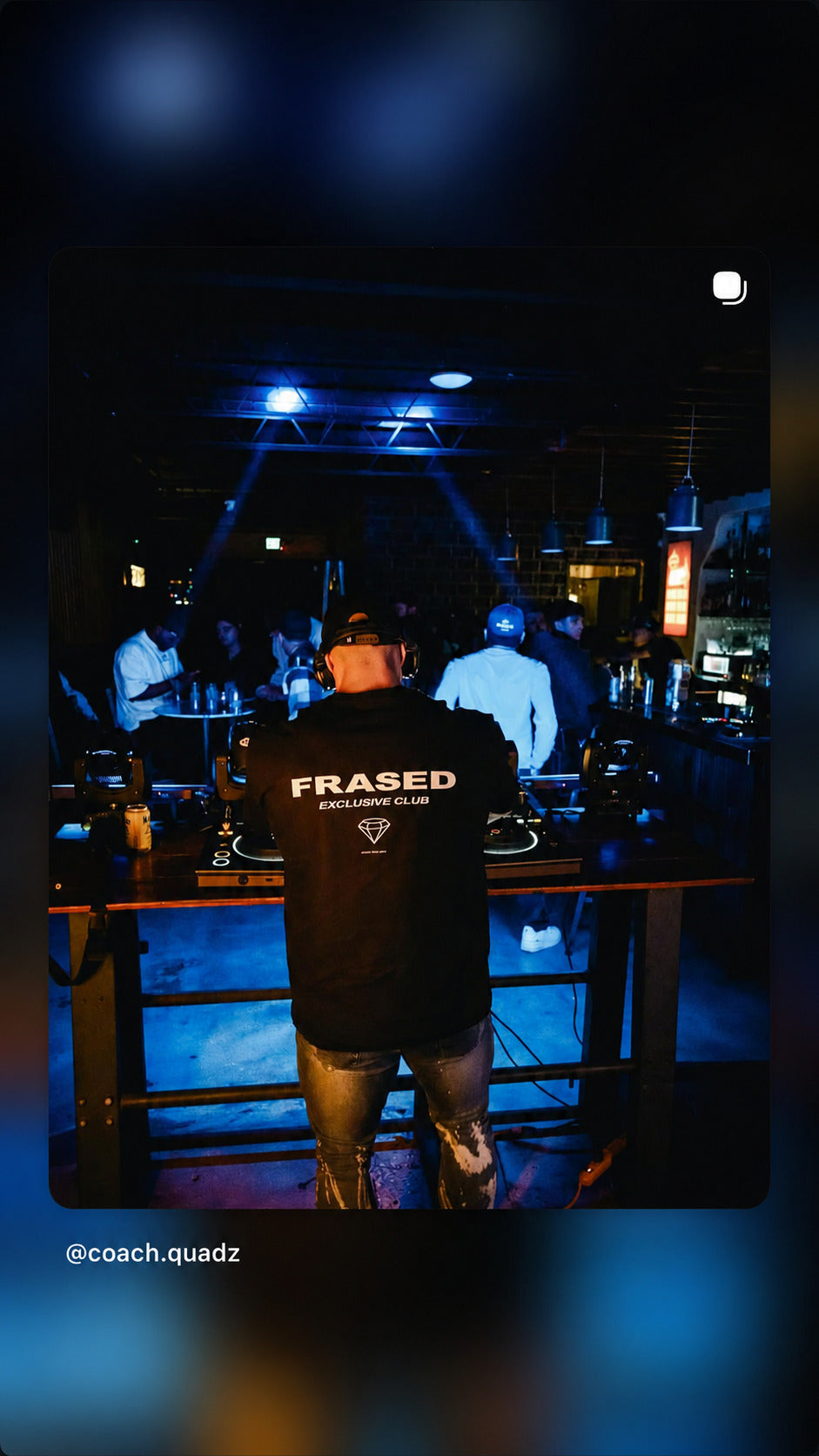DJ at the decks in a dark club wearing a black FRASED Exclusive Club T-shirt with a diamond graphic on the back.