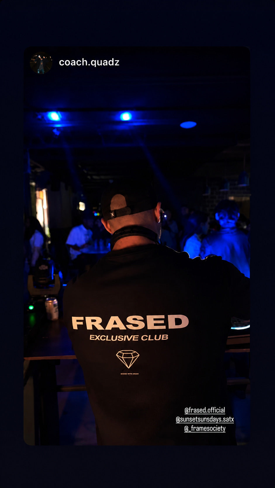 Close-up of a black FRASED Exclusive Club T-shirt with a diamond graphic on the back by a DJ booth.