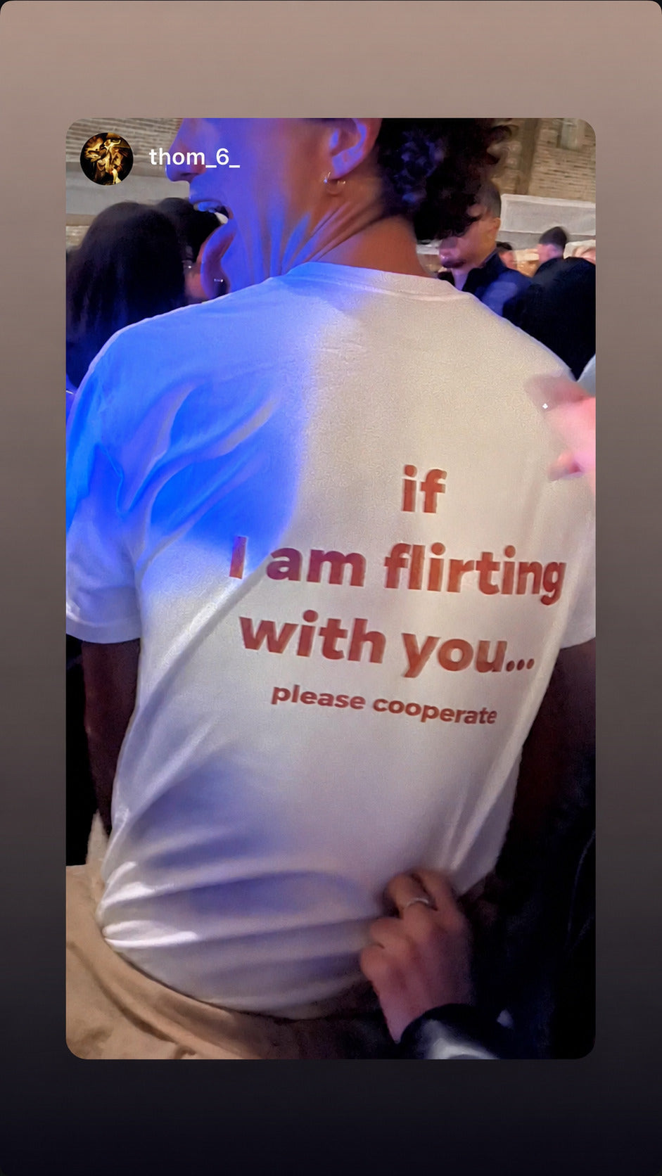 Hvid IF I AM FLIRTING WITH YOU... PLEASE COOPERATE T-shirt set bagfra i en menneskemængde.