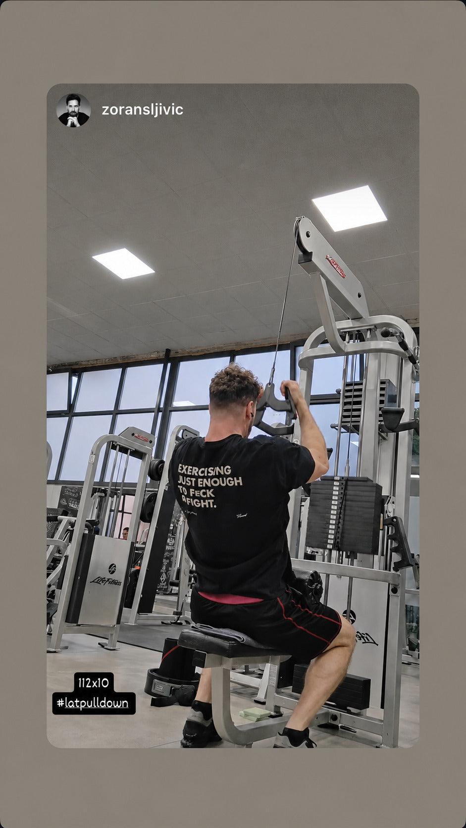 Man exercising at a cable machine in a black “EXERCISING JUST ENOUGH TO F*CK & FIGHT” T-shirt.