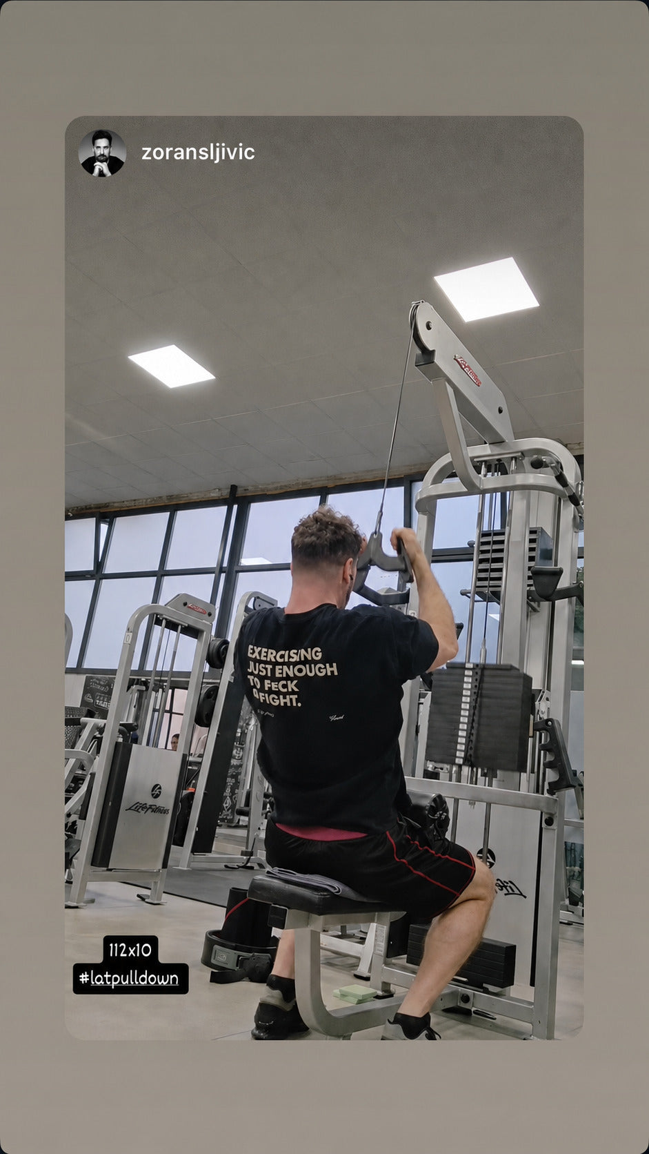 Man exercising at a cable machine in a black “EXERCISING JUST ENOUGH TO F*CK & FIGHT” T-shirt.