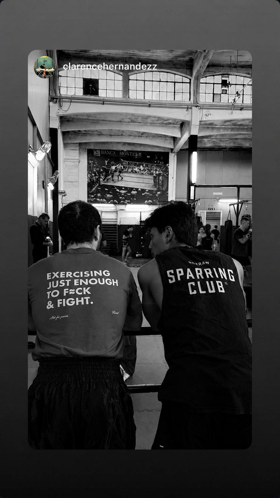 Two people sitting beside a boxing ring; one wears an “EXERCISING JUST ENOUGH TO F*CK & FIGHT” T-shirt.
