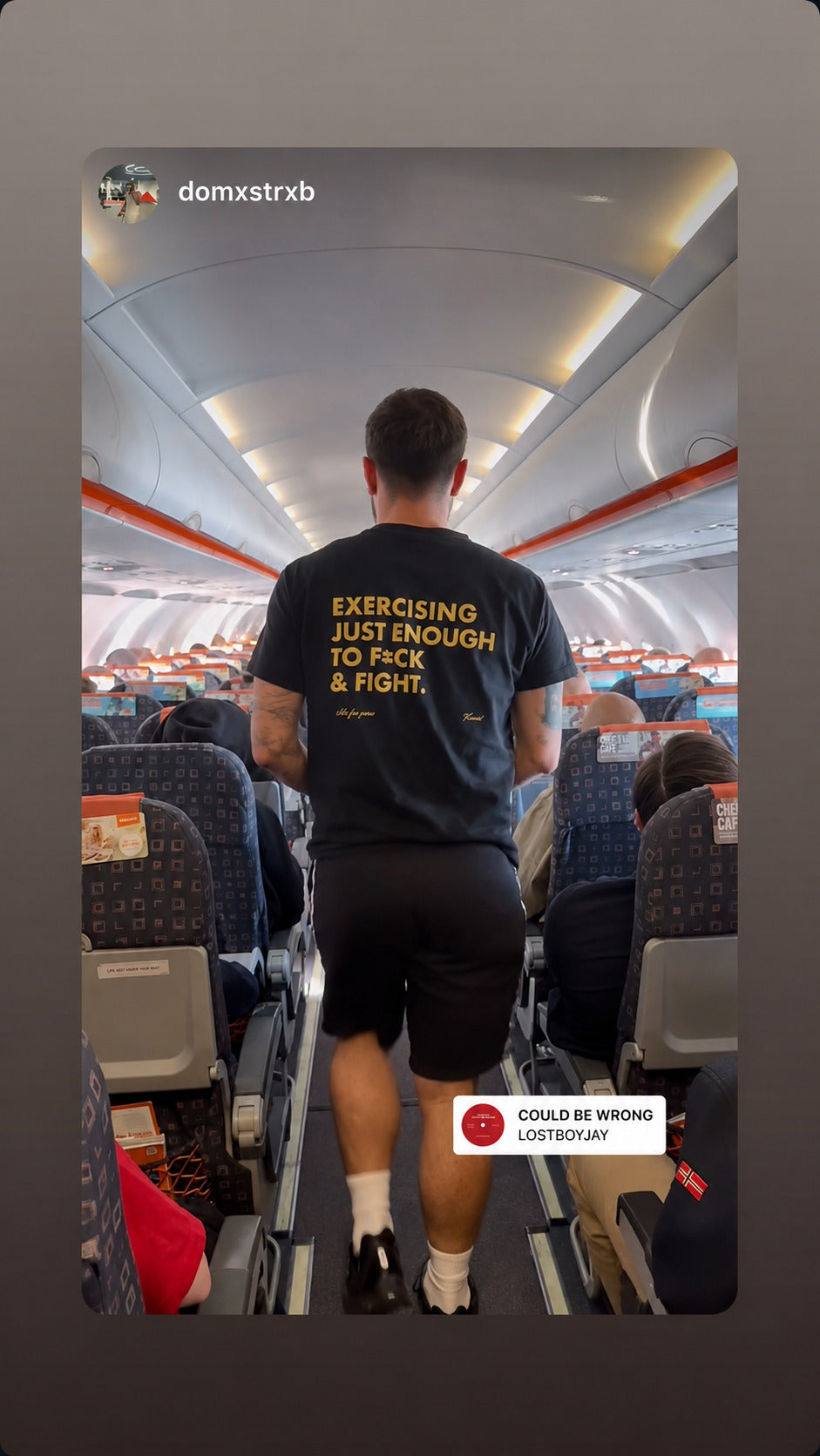 Man walking through an aircraft cabin in a black “EXERCISING JUST ENOUGH TO F*CK & FIGHT” T-shirt.