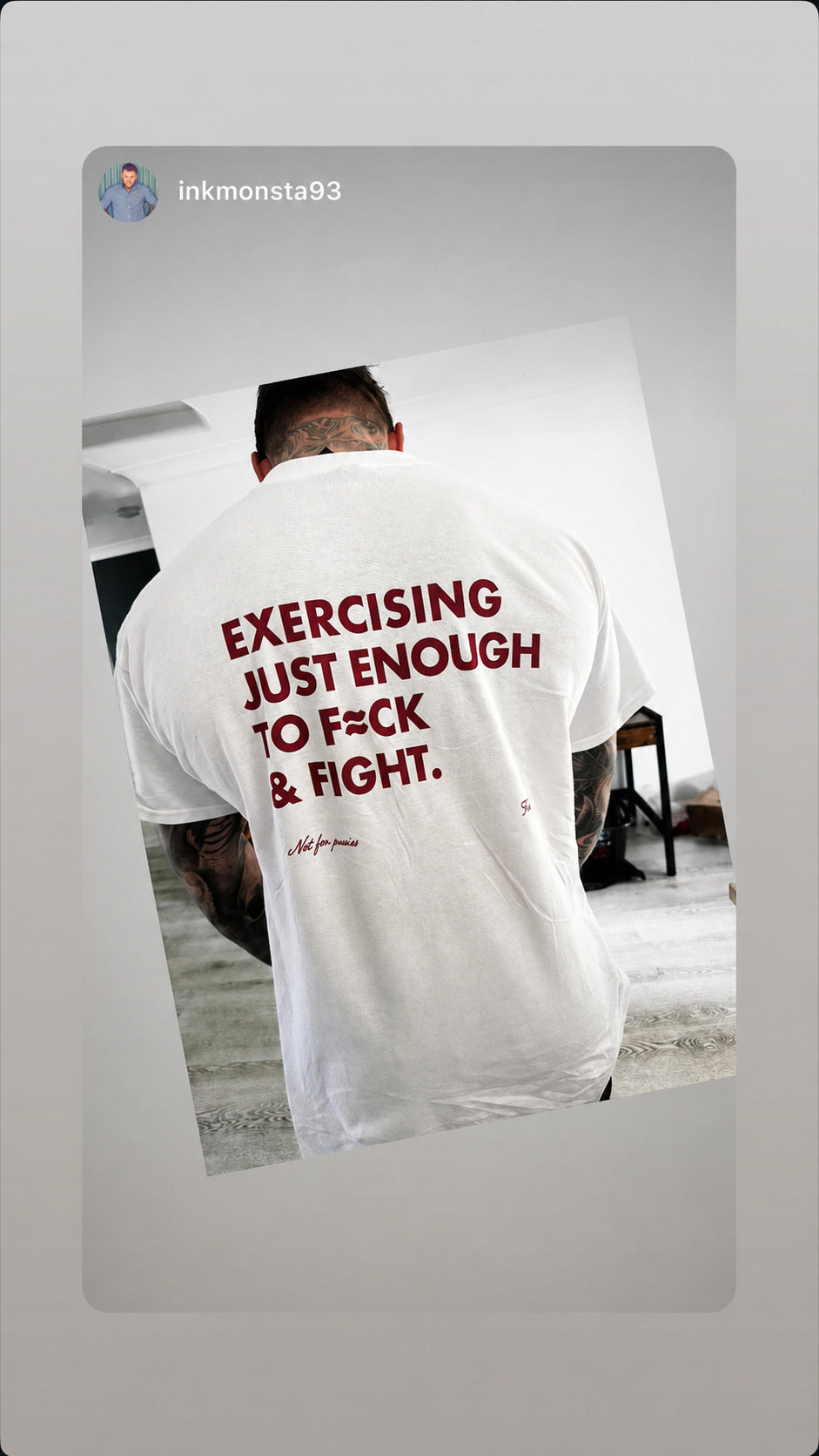 Tattooed man seen from behind in a white “EXERCISING JUST ENOUGH TO F*CK & FIGHT” T-shirt.