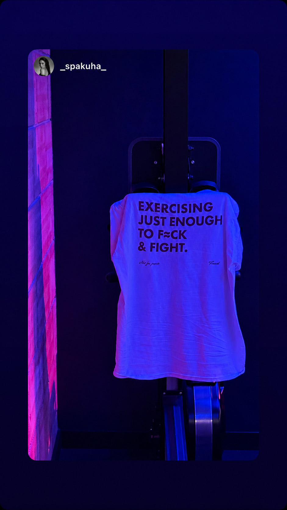 White “EXERCISING JUST ENOUGH TO F*CK & FIGHT” T-shirt hanging under purple and blue lights.