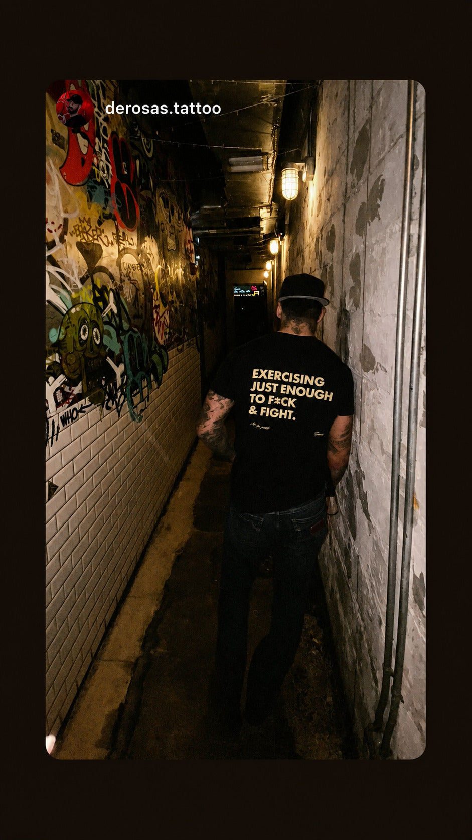 Man in a black “EXERCISING JUST ENOUGH TO F*CK & FIGHT” T-shirt in a narrow graffiti-covered passage.