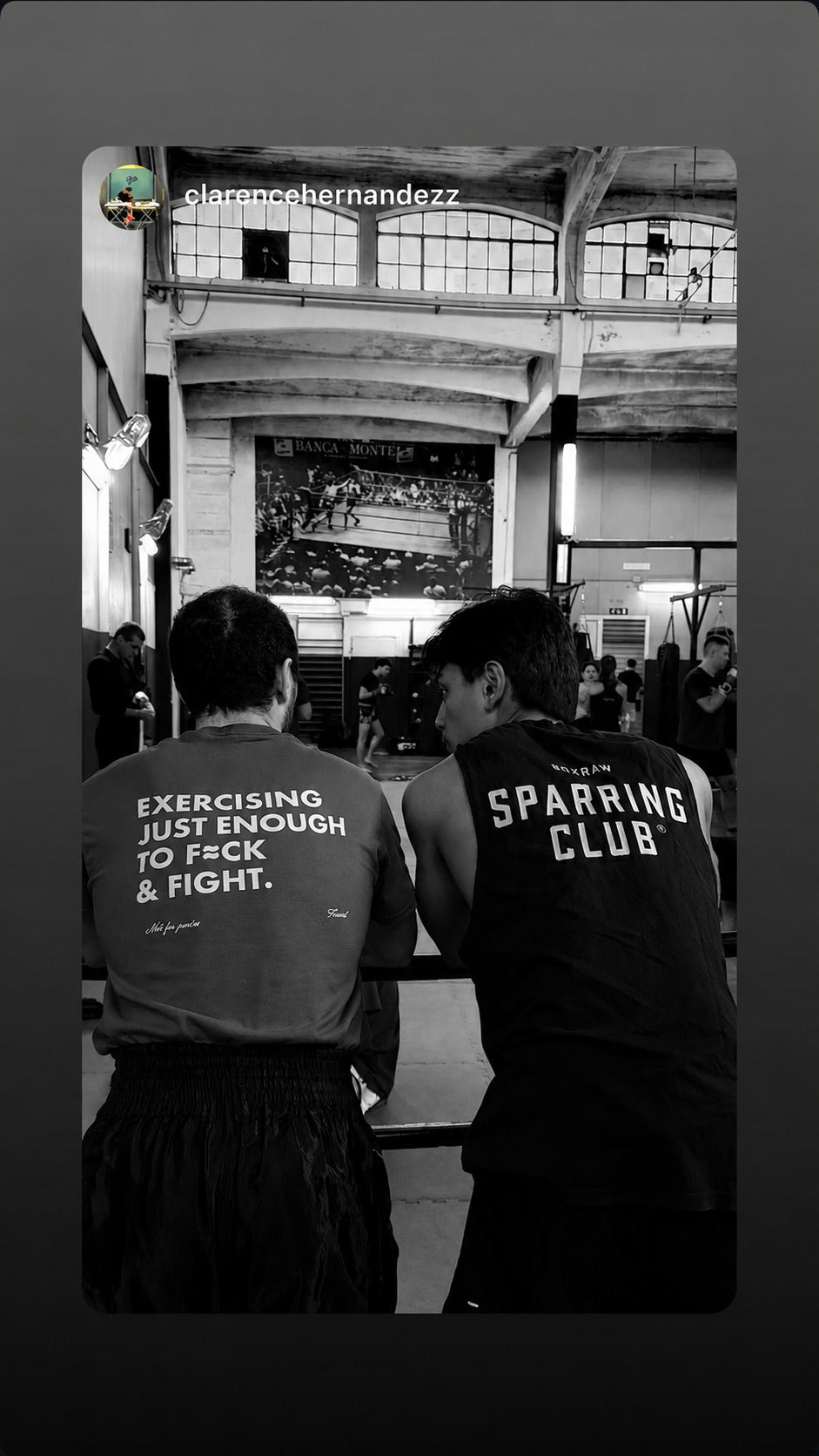 Two people sitting beside a boxing ring; one wears an “EXERCISING JUST ENOUGH TO F*CK & FIGHT” T-shirt.