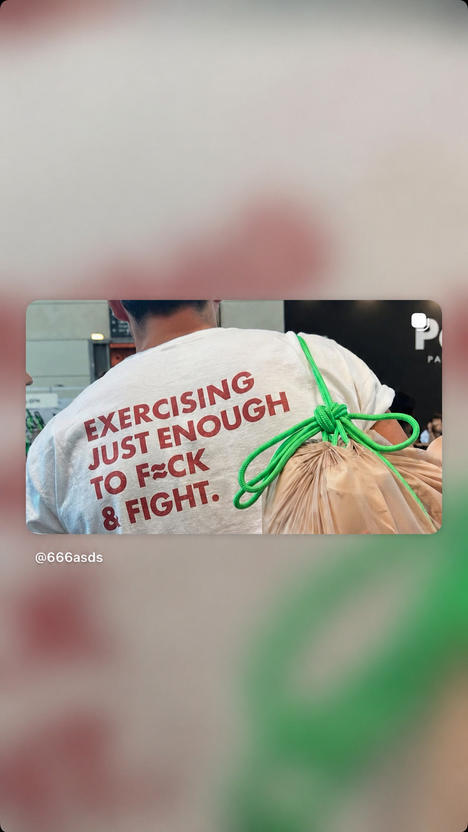 Person in a white “EXERCISING JUST ENOUGH TO F*CK & FIGHT” T-shirt carrying a bag with a green rope.