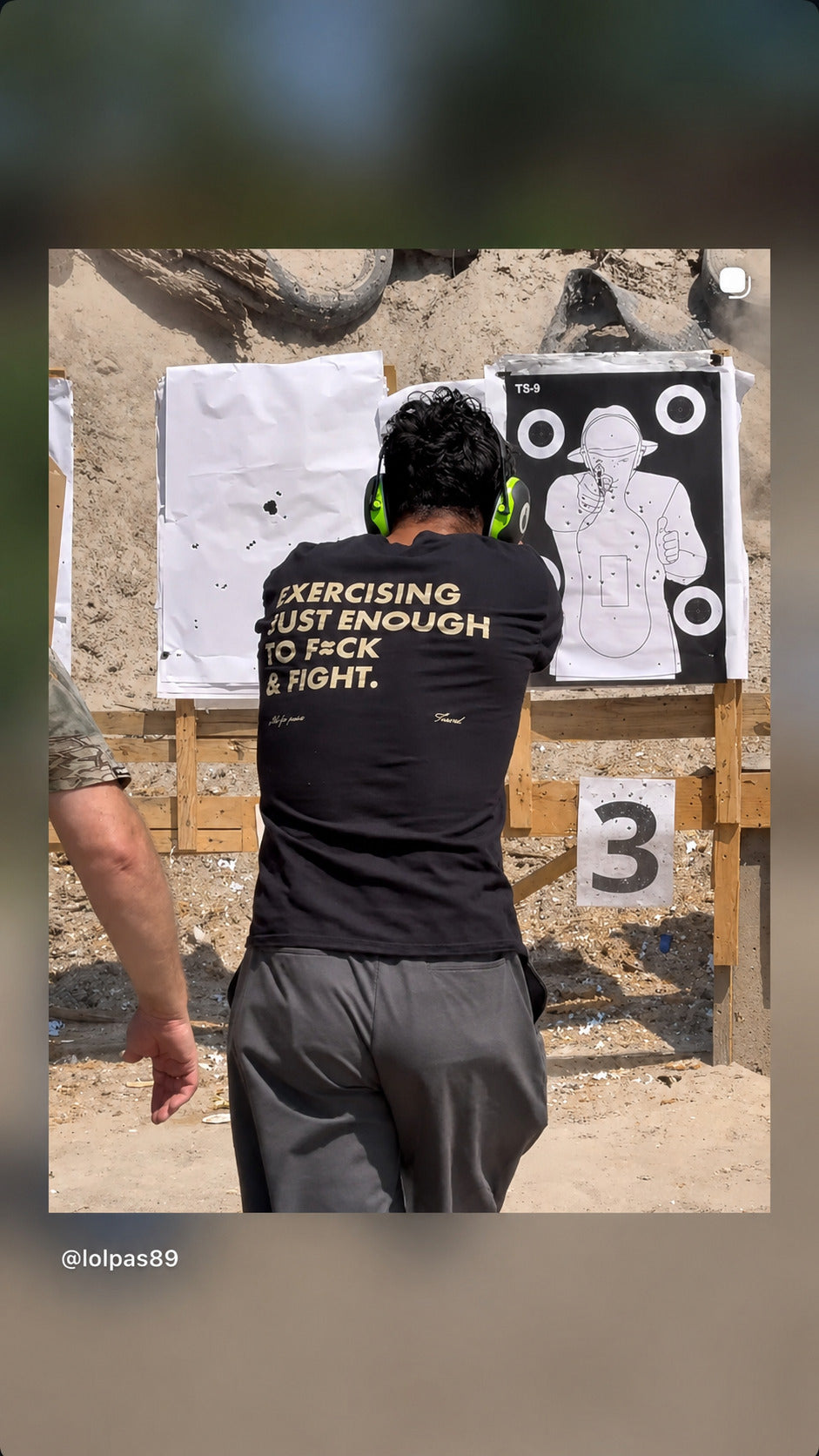 Person by shooting targets in a black “EXERCISING JUST ENOUGH TO F*CK & FIGHT” T-shirt.