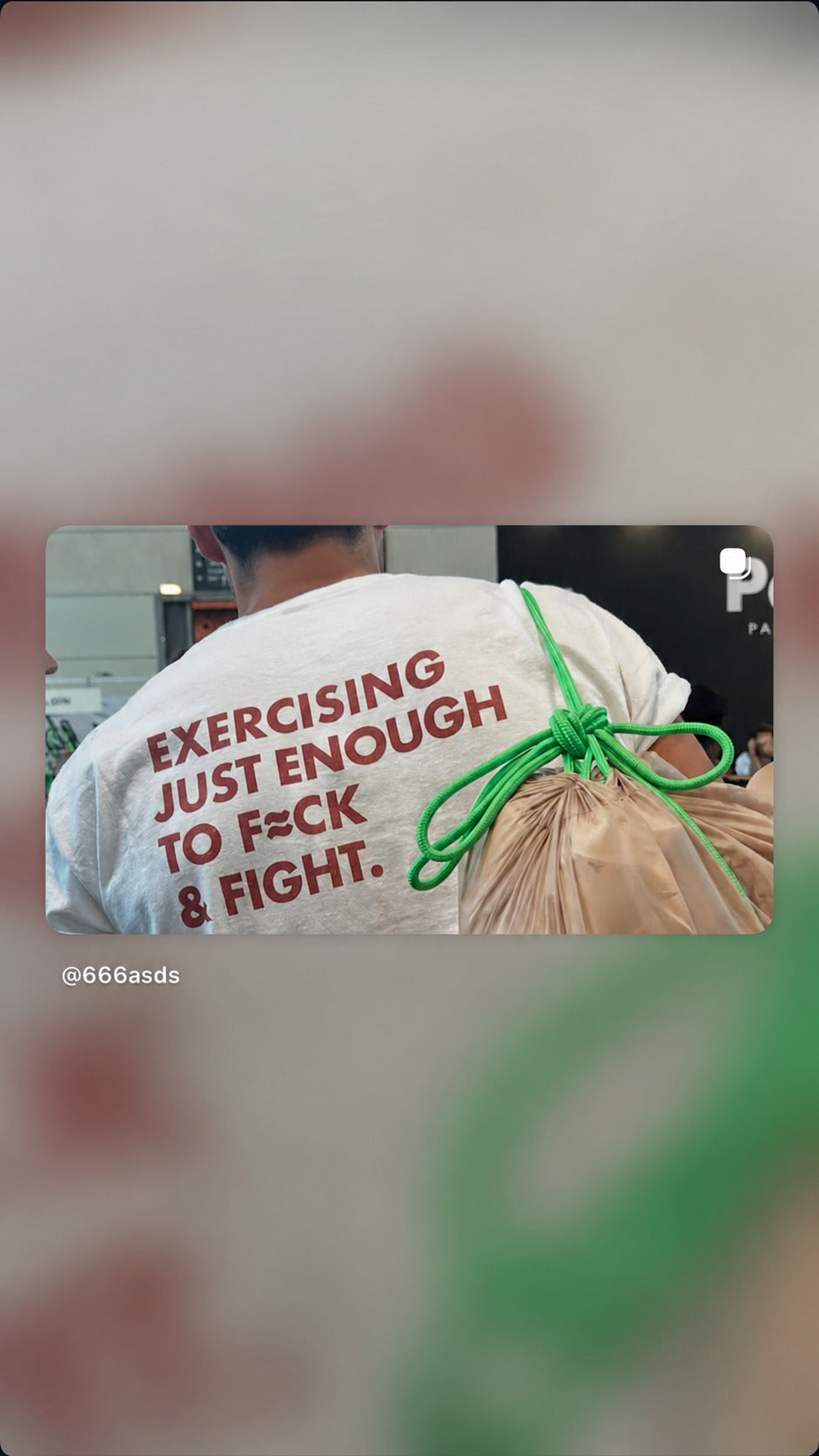 Person in a white “EXERCISING JUST ENOUGH TO F*CK & FIGHT” T-shirt carrying a bag with a green rope.