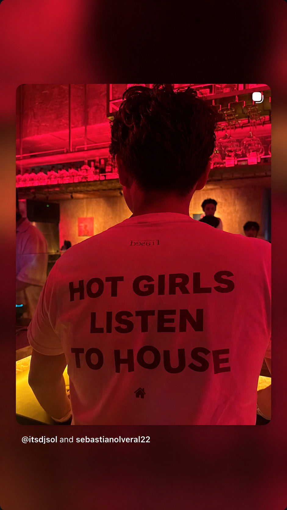 Person seen from behind at a red-lit bar in a white “HOT GIRLS LISTEN TO HOUSE” T-shirt.