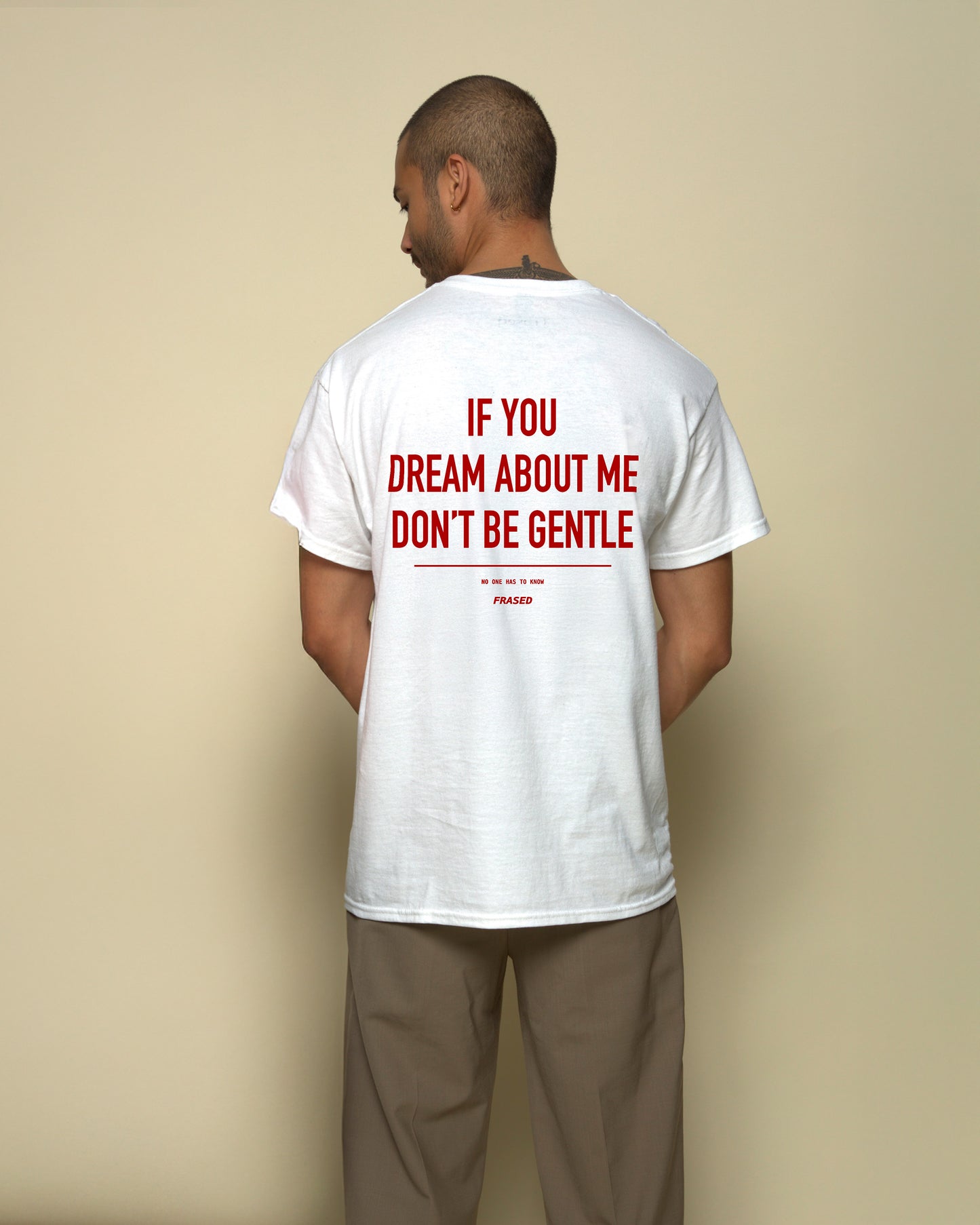 A man viewed from behind wearing a white t-shirt with bold red text that reads 'IF YOU DREAM ABOUT ME DON'T BE GENTLE' and khaki pants, set against a solid beige background.