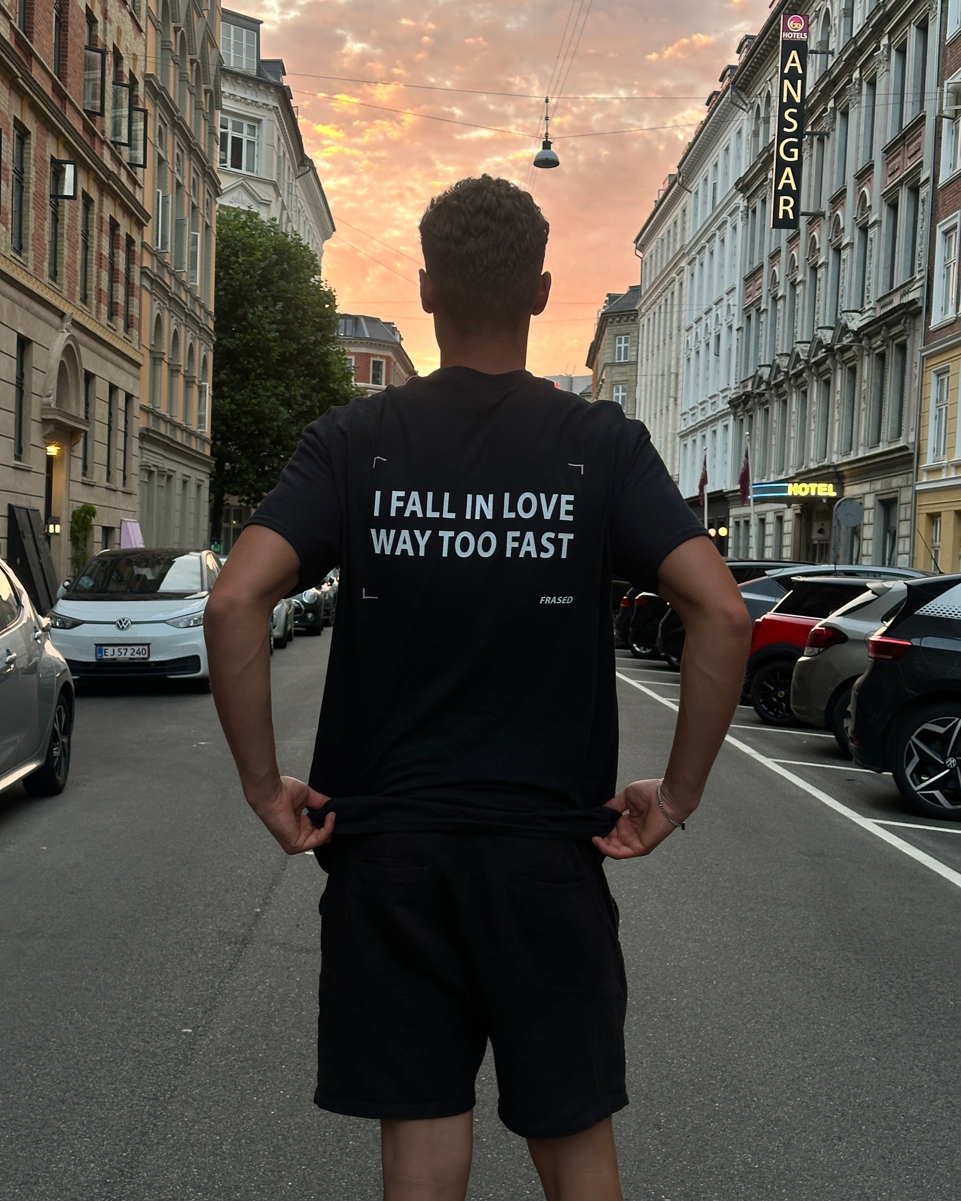 A man wearing a black t-shirt and black shorts stands on a city street, showing the back of the shirt which has white text that reads 'I FALL IN LOVE WAY TOO FAST'. The photo is taken from a low-angle perspective looking up at the buildings and parked cars.