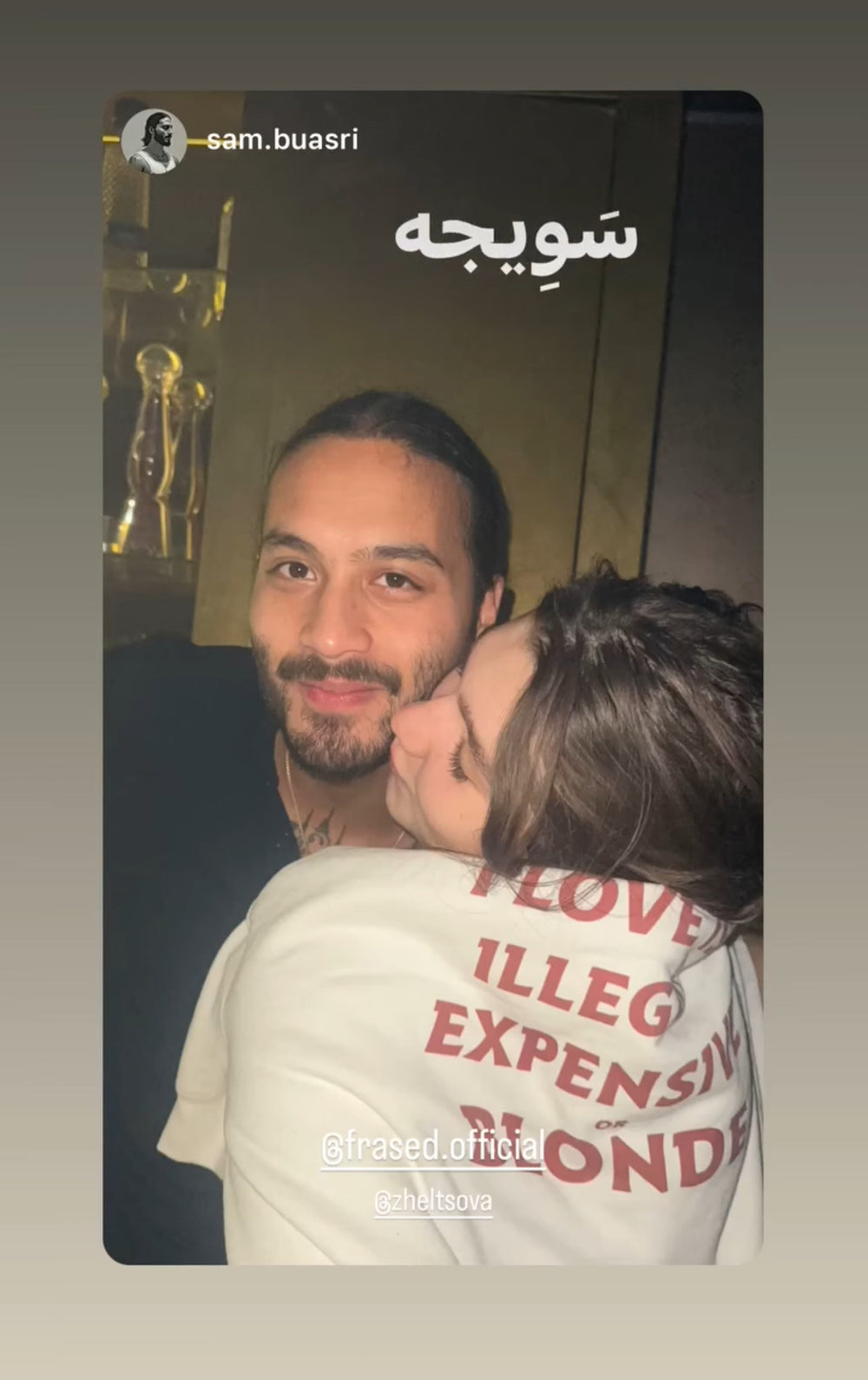 Woman in a white T-shirt with red “EVERYTHING I LOVE IS ILLEGAL EXPENSIVE OR BLONDE” back print hugging a man.