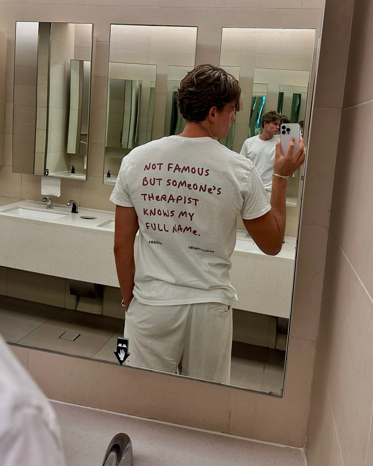 A man takes a mirror selfie in a public bathroom, showing the back of a white t-shirt with red handwritten text that reads 'NOT FAMOUS BUT SOMENATE THERAPIST KNOWS MY FULL NAME.'.