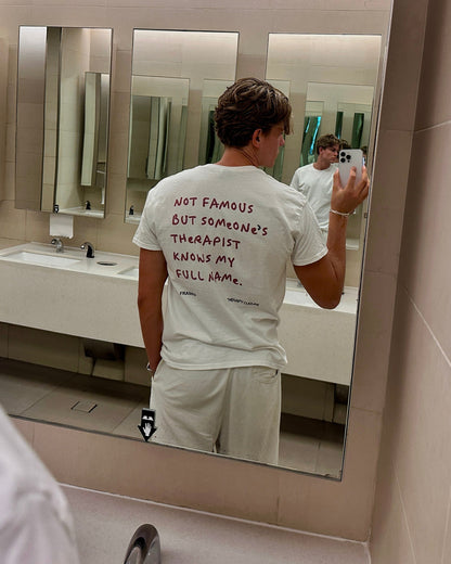 A man takes a mirror selfie in a public bathroom, showing the back of a white t-shirt with red handwritten text that reads 'NOT FAMOUS BUT SOMENATE THERAPIST KNOWS MY FULL NAME.'.