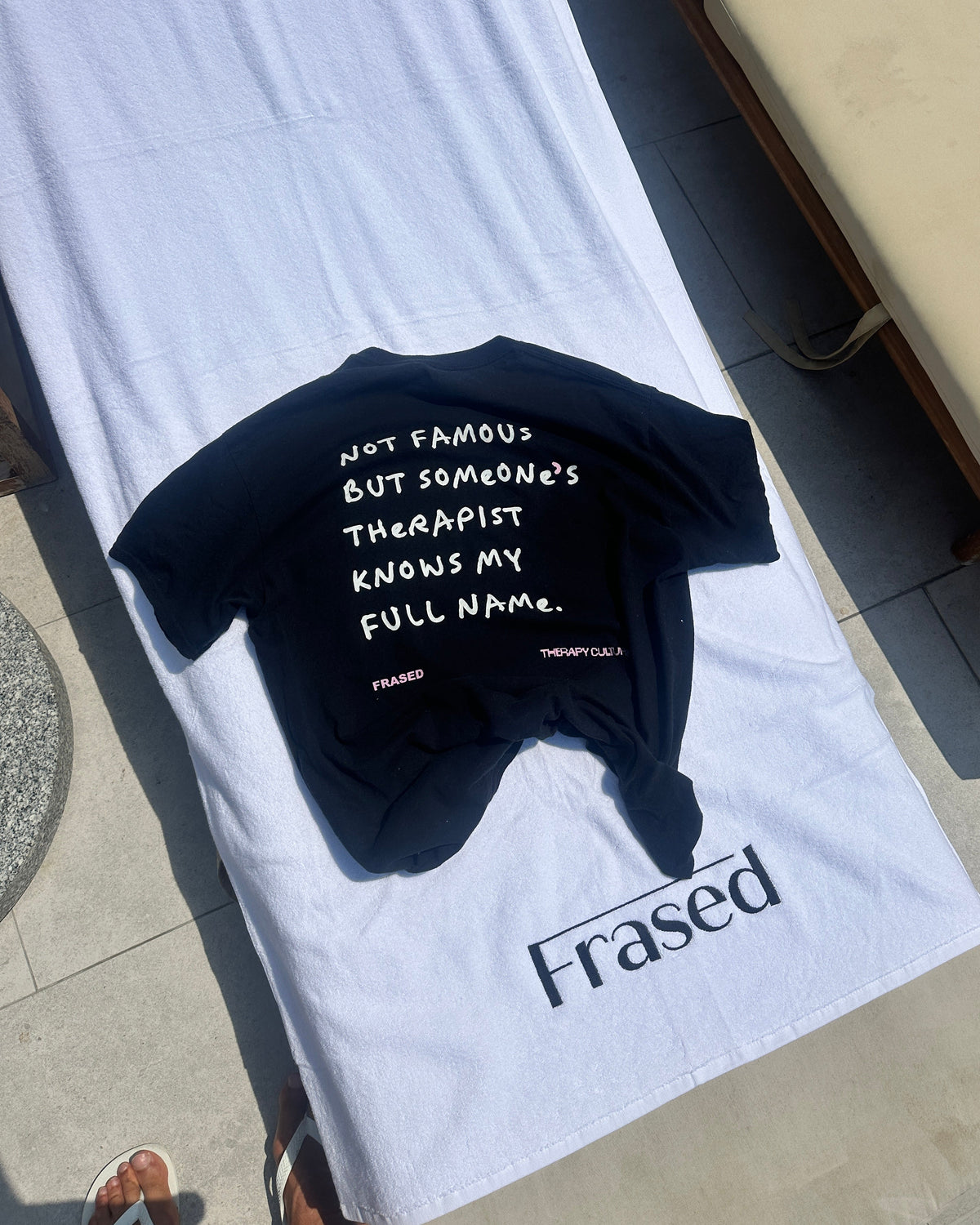 A black short-sleeve T-shirt with white handwritten-style text on the back, laid flat on a white towel outdoors. The towel also has the word 'Frased' printed near the bottom, and a beige lounge chair is visible in the upper right.