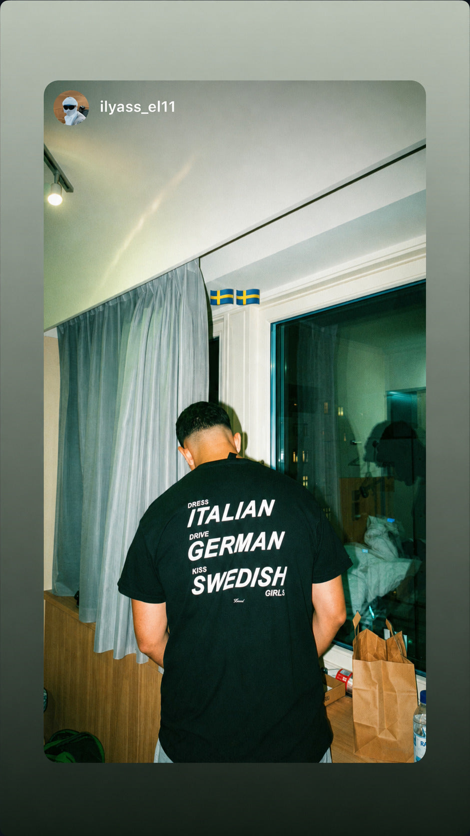 Mand indendørs i sort FRASED T-shirt med hvidt 'Dress Italian. Drive German. Kiss Swedish Girls'-rygtryk.