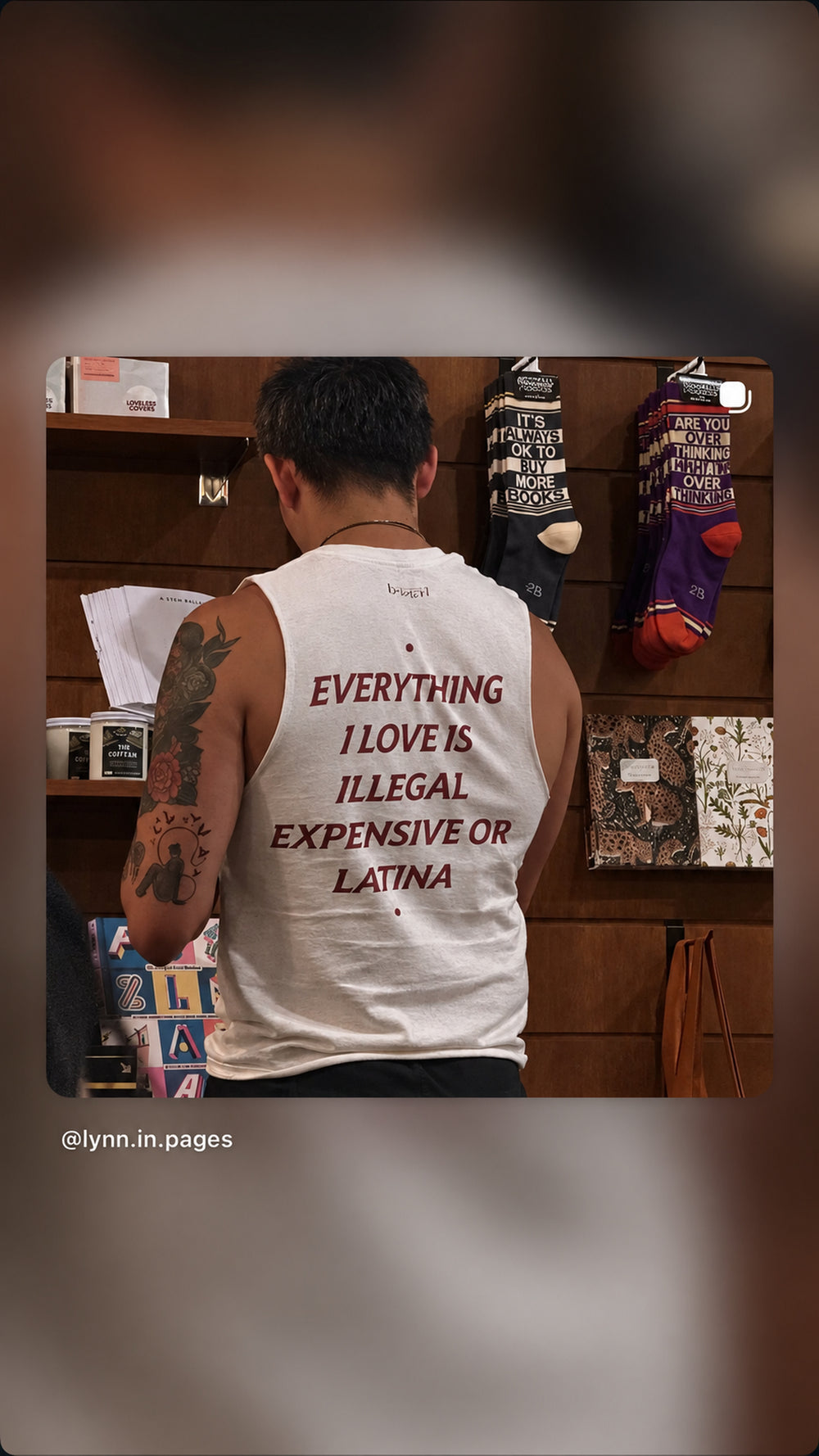 Person i hvid EVERYTHING I LOVE IS ILLEGAL, EXPENSIVE OR LATINA tanktop står ved en hylde.